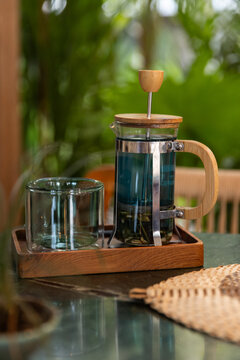 Tea press with butterfly pea flower blue tea served in a tropical cafe with empty tea mug and leafy background. 