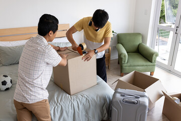 Two men packing cardboard box on bed, preparing for move with suitcase nearby