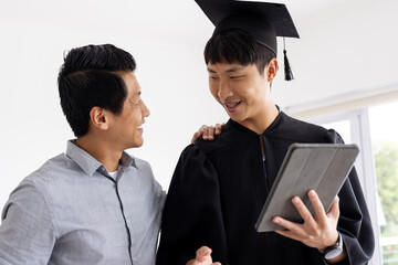Proud father congratulating son in graduation gown holding tablet at home