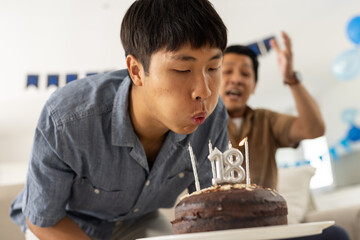 Young man blowing out candles on birthday cake while dad celebrates joyfully