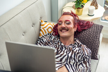 Non-binary person with makeup smiling and using laptop on sofa