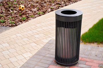 urban trash can on paved pathway with grass and dry leaves in background. nearby, a black and...
