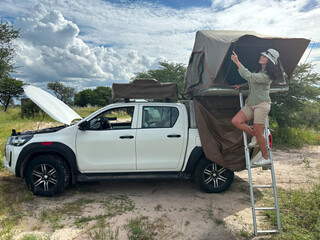A dark-haired woman in safari attire stands on a ladder adjusting a roof tent on an off-road vehicle in the African wilderness. Adventure in the Central Kalahari, Botswana. 