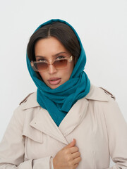 Woman in a beige trench coat with a teal hooded scarf. She wears square sunglasses and gazes...