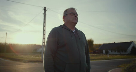 Man standing on rural road at dusk with serene expression, sunset glow in background, rural scene,...