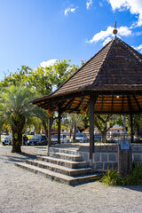 A charming gazebo located in a lush green park, surrounded by trees and bathed in sunlight under a bright blue sky. A peaceful setting perfect for relaxation and leisure.
