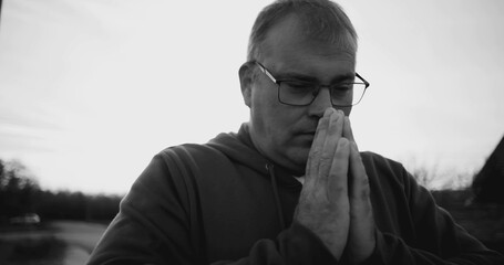 Man with hands clasped in prayer, head bowed, contemplative expression, peaceful rural setting, black and white, spiritual moment, reflection, and gratitude