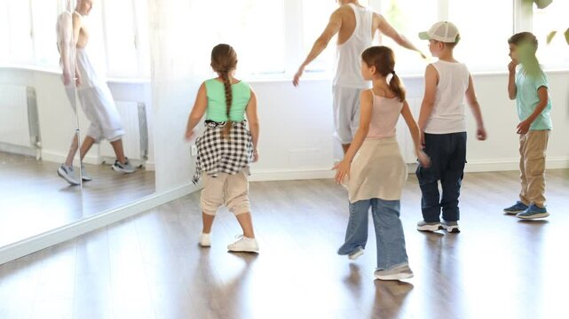 Preteen children enthusiastically following male instructor moves during group hip-hop lesson in sunlit mirrored studio, mastering modern street dance choreography. High quality 4k footage