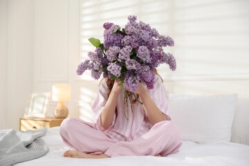 Teenage girl hiding behind bouquet of lilac flowers on bed at home