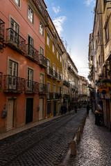 Fototapeta premium A narrow, cobblestone street winds through a historic European district. Vivid, multi-story buildings with wrought iron balconies, tram tracks, and an ATM sign complete this sunlit urban charm
