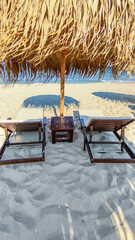 Mobile photo a calm and inviting beach scene with a thatched umbrella and lounge chairs on the sand. The gentle waves and clear sky create a serene and picturesque coastal environment.