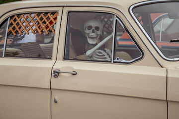 Skeleton driver sits behind the wheel of a vintage beige car, visible through the closed window. The interior includes a latticed design on the door near the window