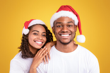 Fototapeta premium Christmas Portrait Of Joyful Black Couple In Santa Hats Hugging Posing Over Yellow Studio Background, Smiling To Camera. Xmas, New Year And Winter Holidays Celebration. Panorama