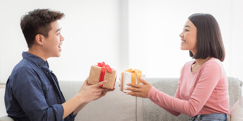Valentine's Day Celebration. Lovely Asian Couple Giving Gifts Boxes Congratulating Each Other Celebrating Holiday Together Sitting On Sofa At Home. Romance, Holidays Presents Concept