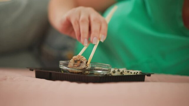 Close up of woman in green top relaxing on bed using chopsticks to lift sushi roll from soy sauce tray, letting excess sauce drip off. Focus on polished nails and calm,
