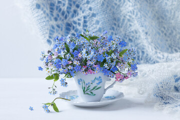 Bouquet of spring flowers forget-me-nots and omphalodes in a cup on the table, lace shawl, blur, beautiful card, calendar