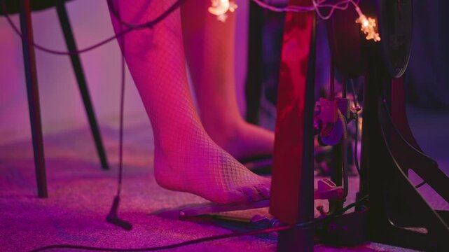Leg view of barefoot woman in fishnet pantyhose stepping on kick pedal during electronic drum practice in softly lit studio with purple glow and visible wiring