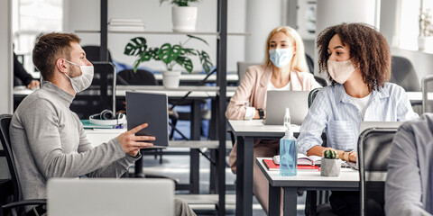 Team of employees working on project and social distancing during outbreak. Millennial male manager in protective mask talk to european and african american women at work desks with laptops