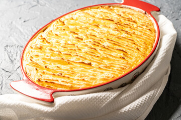 Potato casserole in a porcelain, red baking dish with a white towel on a gray background. Close-up.