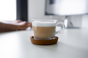 Cappuccino in transparent mug on desk with a male hand and computer monitor in background