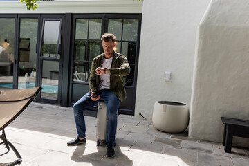 Fototapeta premium Man sitting on suitcase checking time on smartwatch in sunny patio