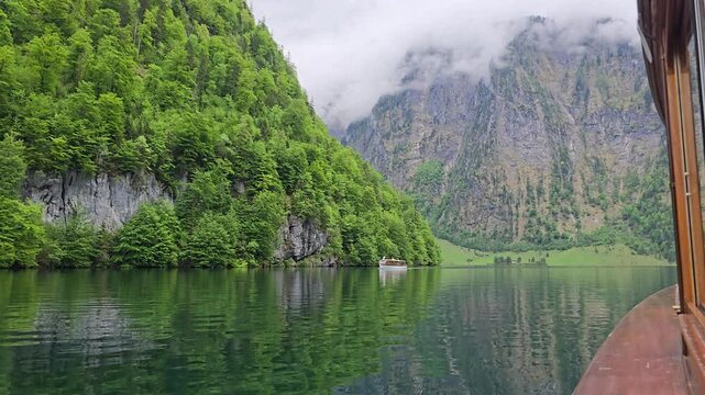A classic ferry glides across the emerald waters of K&ouml;nigssee, framed by lush forests, steep alpine peaks, and drifting clouds in Berchtesgaden National Park, Bavaria.
