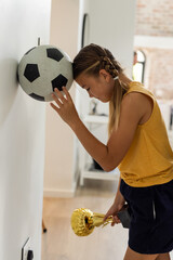 Girl holding trophy, leaning head on soccer ball at home, feeling thoughtful