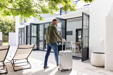 Fototapeta premium Man with suitcase entering modern home through glass doors, ready for travel