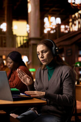 Mature woman attending a webinar from a quiet school library, taking notes during a digital lesson and engaging with her professor and classmates through wireless telecommunication tools.