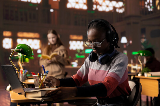 African american girl student listening to a podcast on headset and takes notes, using educational audio and video materials for academic purposes. Scholar prepares for a class assignment.