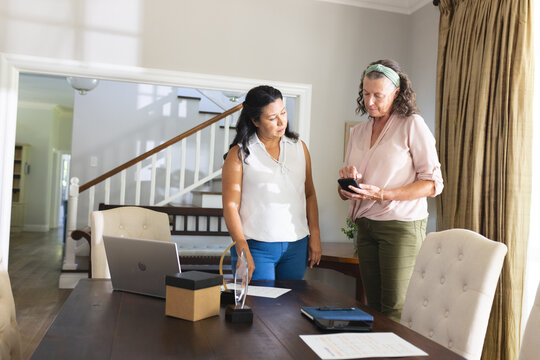 Two women discussing smartphone features at home office with laptop and documents - Powered by Adobe