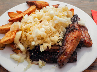 Large meal with potato wedges, well done pork belly slices, and sour white cabbage with onion salad on a plate on a wooden table with red napkin and fork and knife. Dinner made from fine products.