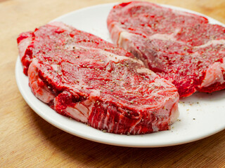 Two premium raw rib eye steaks on a white plate and wooden kitchen table top. Meat is seasoned with pepper and herbs. Cooking luxurious meat product concept.