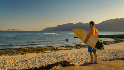 Male surfer stands barefoot on beach gazing at the small rolling waves. Young man holds his wooden surfboard, checking out surf spot in morning light and deciding whether to go for a surf session.