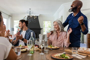 Friends celebrating at dining table, raising glasses and clapping joyfully indoors