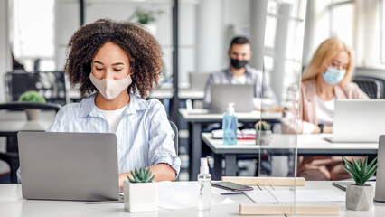 Fototapeta premium Modern devices for working in coworking office during virus outbreak. African american woman in protective mask typing on laptop at table with smartphone, antiseptic and protective glass, empty space