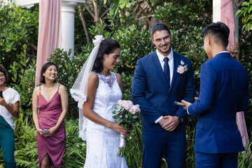 Bride holding bouquet, smiling at outdoor wedding with groom and officiant present