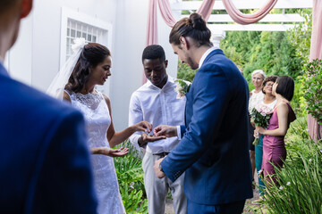 Bride and groom exchanging rings outdoors, surrounded by friends and family