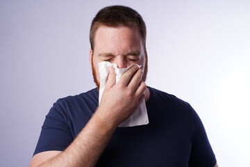 Young guy with ginger beard blows his nose in a tissue.