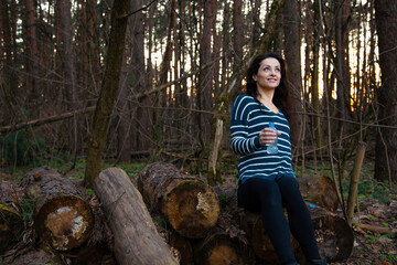 Beautiful happy emotion woman hiking in the forest relax sitting on logs looking up on sunny spring weather. Girl rest enjoy, drinking water from plastic bottle in style fashion clothes in woods