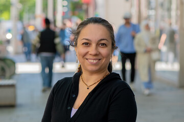 A smiling beautiful woman standing against a backdrop of city buildings on a sunny day.