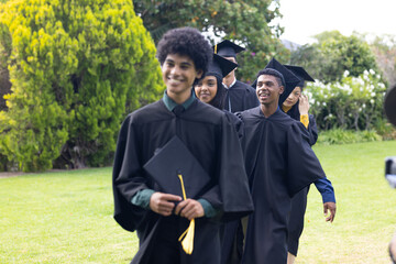 Graduating students in caps and gowns walking joyfully on green lawn