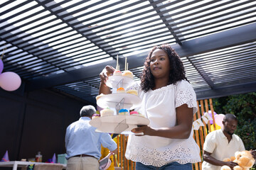 African American woman arranging colorful cupcakes on tiered stand at family party