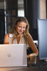 Businesswoman working late at office, smiling while using laptop with coffee nearby