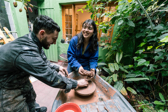 Two smiling ceramists working with raw clay, preparing it before using the pottery wheel in their workshop, surrounded by plants - Powered by Adobe