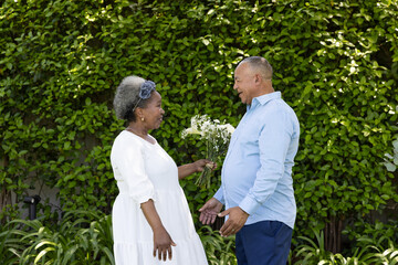 African American senior couple exchanging flowers and smiling in lush garden