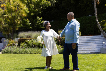 Naklejka premium Senior couple holding hands and smiling while walking together in sunny garden