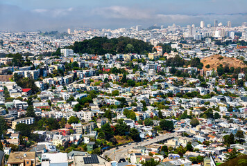Densely packed residential neighborhoods of San Francisco viewed from above, showing colorful houses on steep hills, Sutro Forest in the middle distance, and a fog-covered skyline in the background on