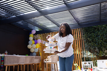 African American woman arranging cupcakes on stand under backyard pergola, with balloon garland