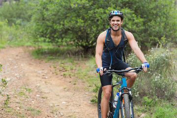 Pedaling man riding mountain bike on narrow trail in woods, with hydration pack, helmet, copy space